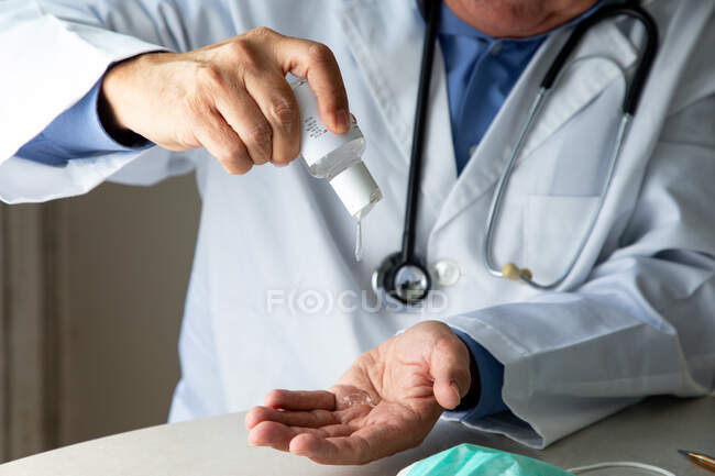 Terapeuta masculino em vestido médico sentado à mesa no hospital e higienizando as mãos com anti-séptico — Fotografia de Stock