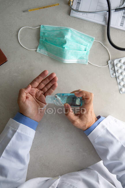 Male therapist in medical gown sitting at table in hospital and sanitizing hands with antiseptic — Stock Photo