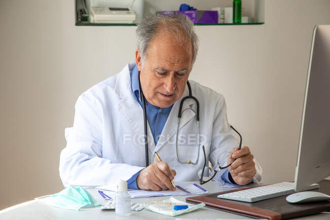 Medico generico di sesso maschile in abito medico seduto a tavola e relazione scritta — Foto stock