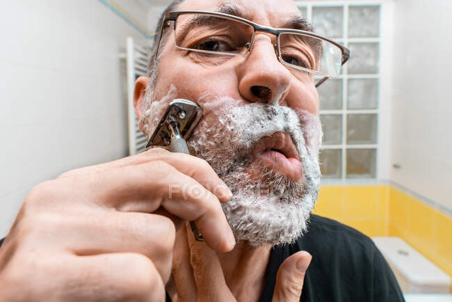 Бородатый мужчина в очках бреется в ванной комнате — стоковое фото