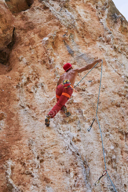 Dal basso di forte alpinista barbuto senza maglietta in berretto rosso arrampicata salita pendio roccioso di montagna in giornata soleggiata senza nuvole — Foto stock