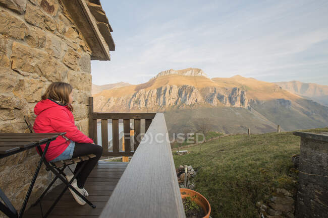 Vista lateral de la chica en ropa casual sentada en silla en la terraza de madera de la casa de piedra en España y admirando el pintoresco paisaje de montaña mientras mira hacia otro lado - foto de stock