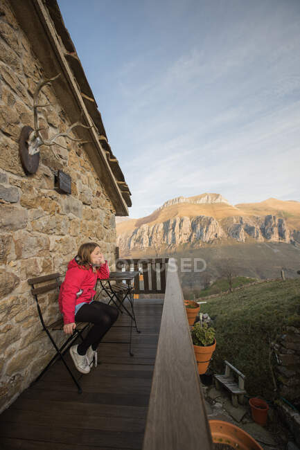 Vista lateral de la chica en ropa casual sentada en silla en la terraza de madera de la casa de piedra en España y admirando el pintoresco paisaje de montaña mientras mira hacia otro lado - foto de stock