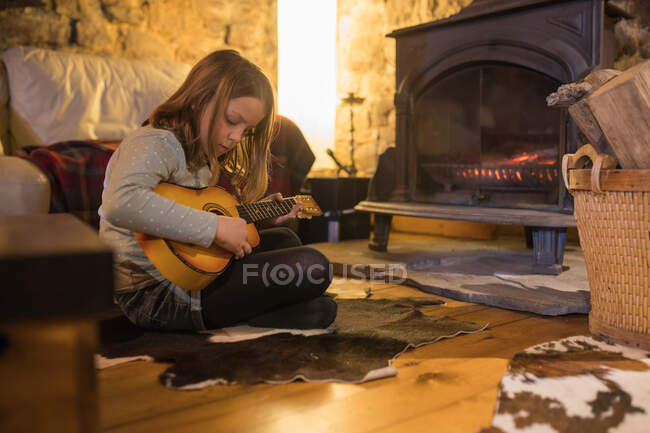 Chica adolescente enfocada en ropa casual sentada con las piernas cruzadas en cómodo sofá y jugando ukelele en casa de piedra durante las vacaciones en Cantabria - foto de stock