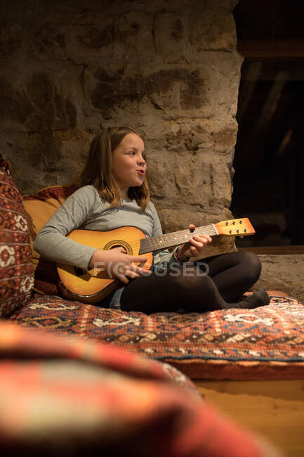 Adolescente ragazza in abiti casual seduta con gambe incrociate su comodo divano e giocare ukulele in casa di pietra durante le vacanze in Cantabria — Foto stock
