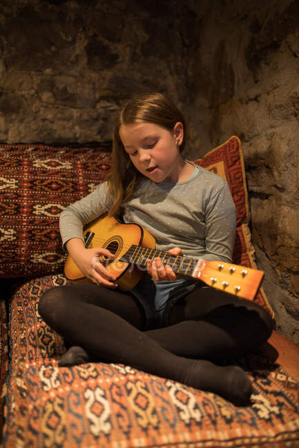 Menina adolescente em roupas casuais sentado com pernas cruzadas no sofá confortável e jogando ukulele em casa de pedra durante as férias na Cantábria — Fotografia de Stock