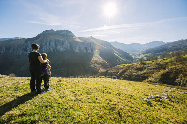 Vista posterior de una excursionista anónima abrazando a los niños mientras está de pie en el prado verde y observa el pintoresco paisaje de las montañas en un día soleado en Cantabria - foto de stock