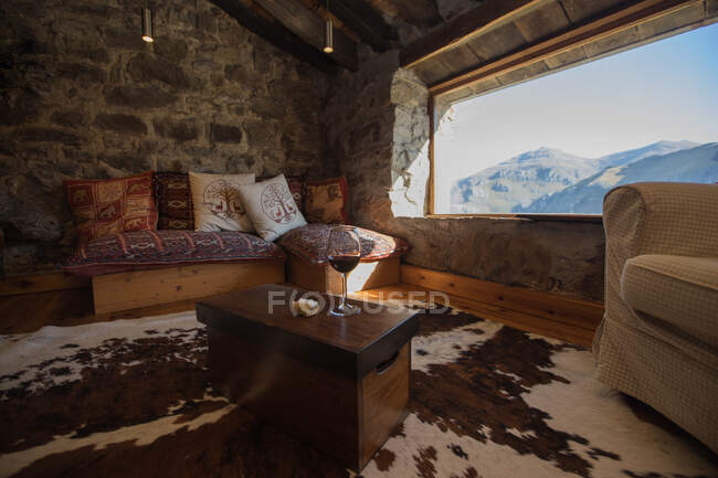Interior de acogedora casa de campo en Cantabria con cómodo sofá con cojín de colores y copa de vino tinto en la mesa frente a una amplia ventana - foto de stock