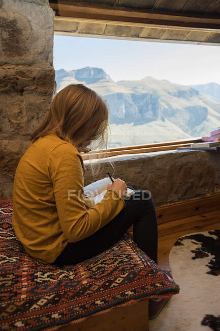 Vista lateral de la chica en ropa casual sentado en un acogedor sofá con alfombra cerca de la ventana en la antigua casa de piedra y tomando notas en copybook - foto de stock