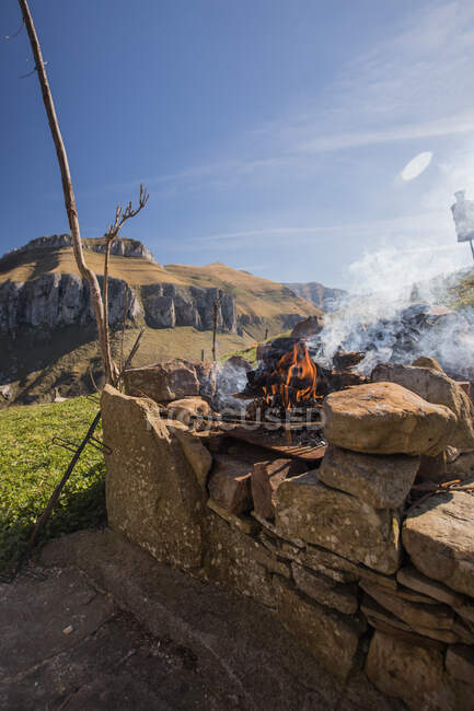 Fumar fuego con llamas en la chimenea de piedra durante el camping en el campo de las montañas de Cantabria en el día soleado - foto de stock