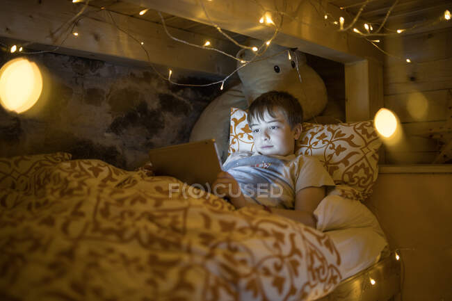 Zufriedener Junge liegt in gemütlichem Bett und benutzt Tablette, während er sich im Schlafzimmer ausruht, das mit glühender, gemütlicher Girlande dekoriert ist — Stockfoto