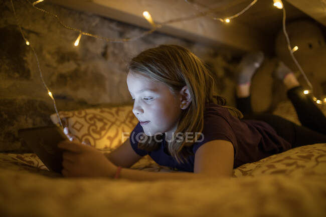 Charmante adolescente en vêtements décontractés se refroidissant sur un lit confortable sous guirlande rayonnante et tablette de navigation dans les loisirs — Photo de stock