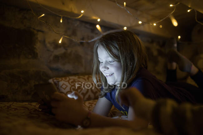 Charming teen girl in casual clothes chilling on cozy bed under glowing garland and browsing tablet in leisure — Stock Photo