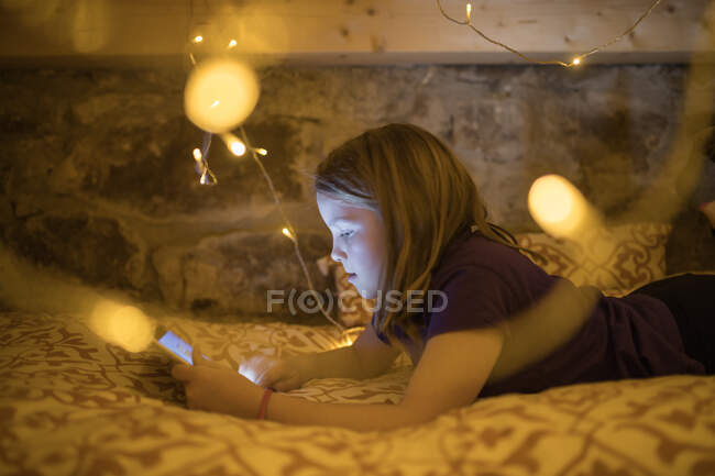 Очаровательная девушка-подросток в повседневной одежде отдыхает на уютной кровати под светящейся гирляндой и просматривает планшет в свободное время — стоковое фото