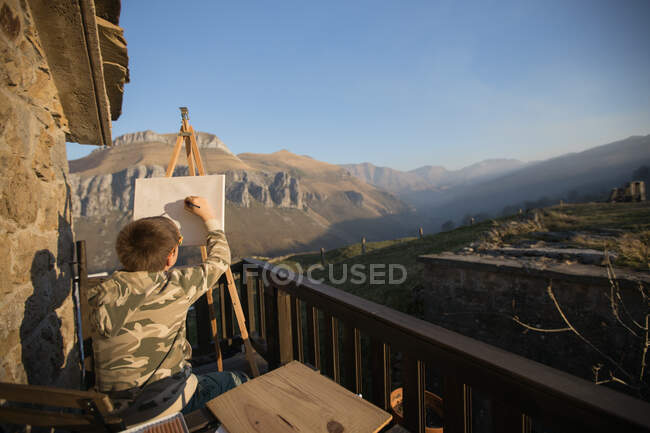 Visão traseira do menino sem rosto sentado no cavalete na varanda pitoresca da casa de campo e desenho na Cantábria — Fotografia de Stock