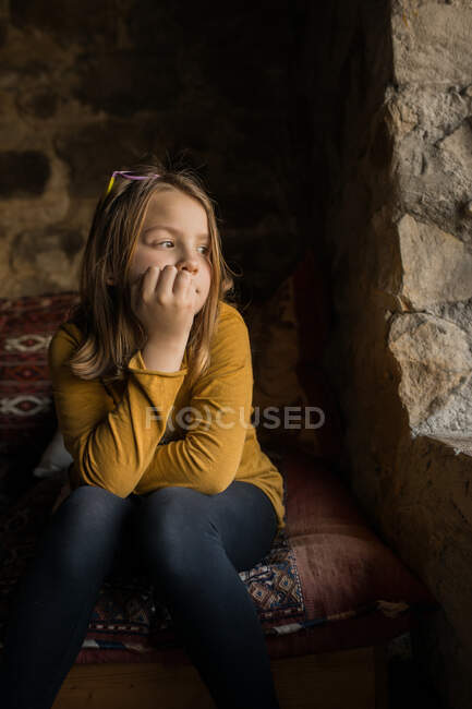 Menina solitário pensativo em roupas casuais inclinando-se na mão enquanto sentado no sofá e olhando para longe na janela da antiga casa de pedra na Espanha — Fotografia de Stock