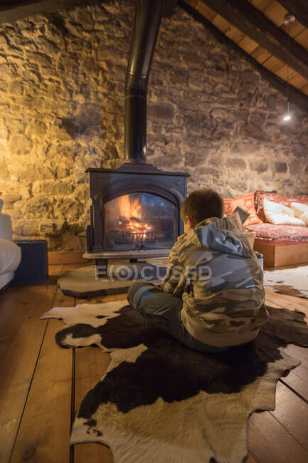 Back view of kid in casual clothes sitting on wooden floor against burning fireplace in cozy room of stone house in Spain — Stock Photo