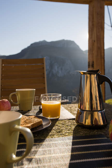 Белые чашки и горячая кофеварка помещены на стол с горячим тостом и стаканом свежего апельсинового сока на открытой веранде с горы на заднем плане — стоковое фото