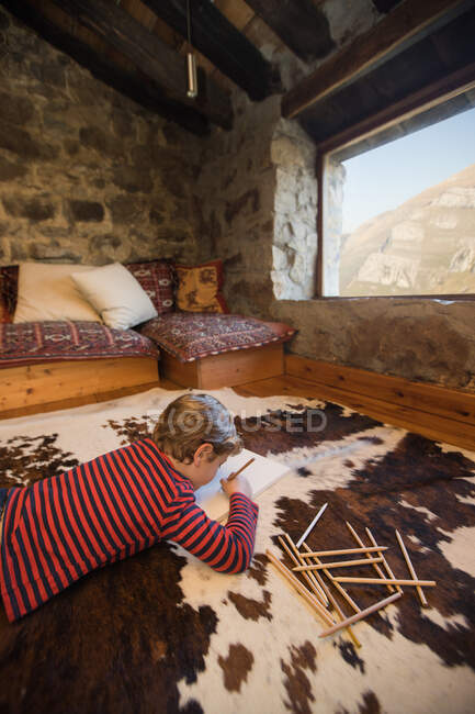 Niño acostado en el suelo sobre una alfombra acogedora y dibujo con lápices de colores en cuaderno de bocetos escalofriante acogedor salón de casa de piedra en Cantabria - foto de stock