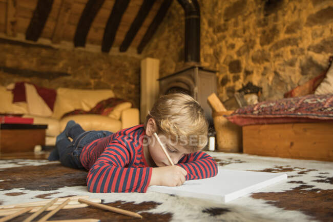 Junge auf dem Boden liegend auf gemütlichem Teppich und Zeichnung mit Buntstiften in Skizzenbuch kühlt gemütliches Wohnzimmer des Steinhauses in Kantabrien — Stockfoto