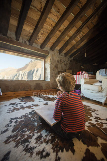 Мальчик сидит на полу на уютном ковре и рисует цветными карандашами в альбоме эскизов, охлаждая уютную гостиную каменного дома в Кантабрии — стоковое фото