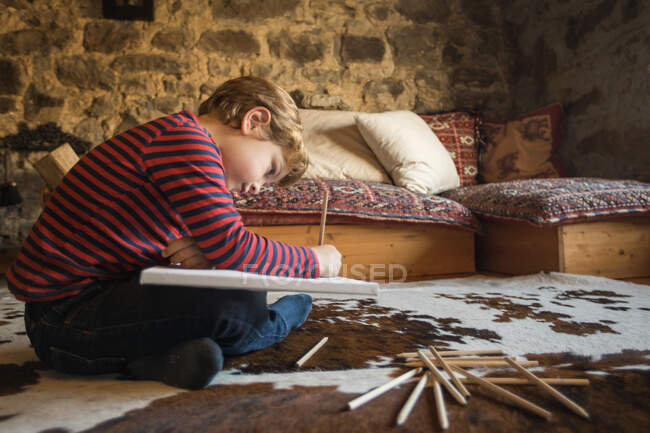 Junge sitzt auf dem Boden auf gemütlichen Teppich und zeichnet mit Buntstiften in Skizzenbuch kühlen gemütlichen Wohnzimmer des Steinhauses in Kantabrien — Stockfoto