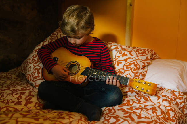 Сконцентрована дитина в повсякденному одязі, сидить на зручному ліжку в спальні і грає на укулеле під час охолодження вдома в Кантабрії. — стокове фото
