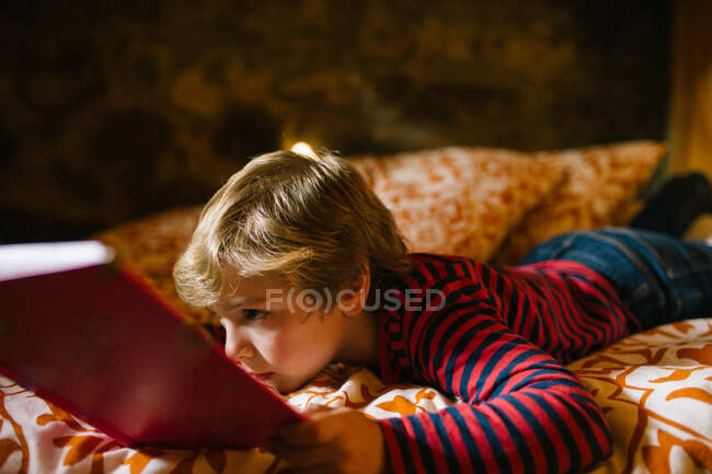 Niño de ensueño en ropa casual acostado en la cama acogedora y disfrutando de un interesante cuento de hadas en el libro de cuentos para niños - foto de stock