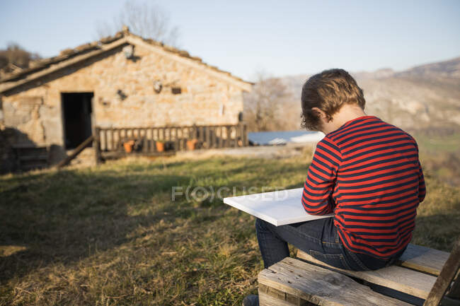 Back view of adorable child sitting on wooden bench and painting on canvas during weekend on background of magnificent mountainous landscape — Stock Photo