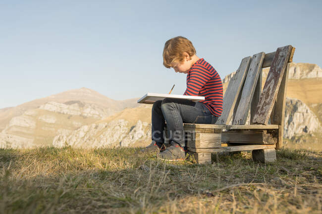 Side view of adorable child sitting on wooden bench and painting on canvas during weekend on background of magnificent mountainous landscape — Stock Photo