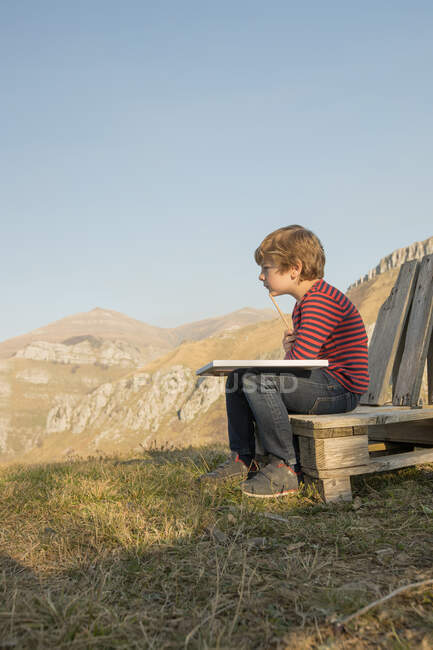Vue latérale de l'adorable enfant assis sur un banc en bois et peinture sur toile pendant le week-end sur fond de magnifique paysage montagneux — Photo de stock