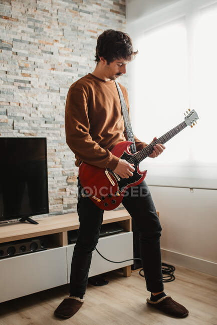 Homem adulto de corpo inteiro tocando guitarra elétrica em pé no quarto aconchegante em casa — Fotografia de Stock