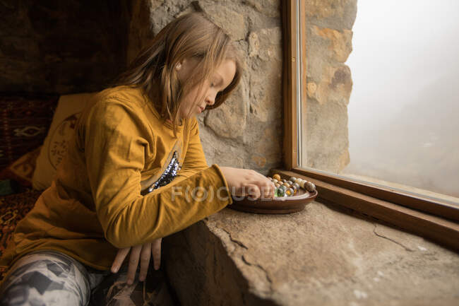 Side view of calm kid in casual clothes sitting on bed at stone windowsill in country house and entertaining with colorful glass marbles — Stock Photo