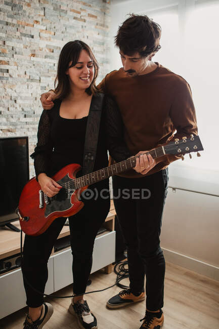Adult guy with mustache teaching smiling girlfriend to play electric guitar while spending time at home together — Stock Photo
