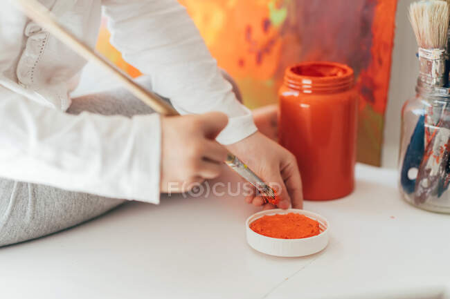 Kreative blonde Mädchen in lässiger Kleidung sitzen auf Fensterbank gegen Fenster und malen mit Pinsel großen mehrfarbigen Regenbogen auf orangefarbener Leinwand — Stockfoto
