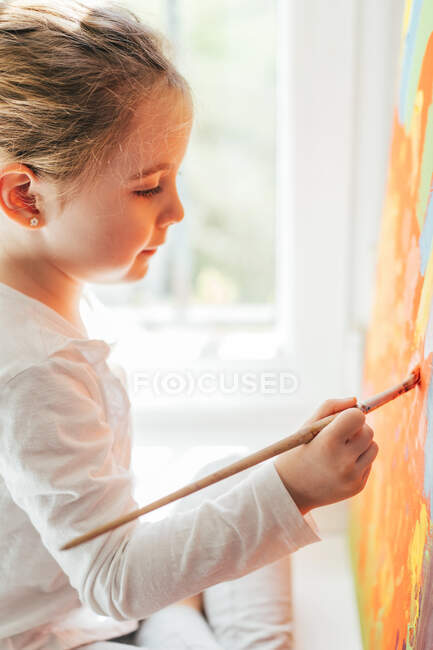 Creative blond girl in casual clothes sitting on window sill against window and painting with paintbrush large multi colored rainbow on orange canvas — Stock Photo