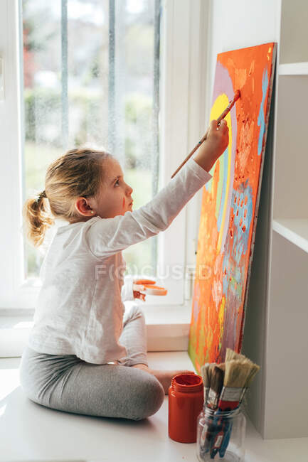 Творческая блондинка в повседневной одежде сидит на подоконнике у окна и рисует кисточкой большой разноцветной радугой на оранжевом холсте — стоковое фото