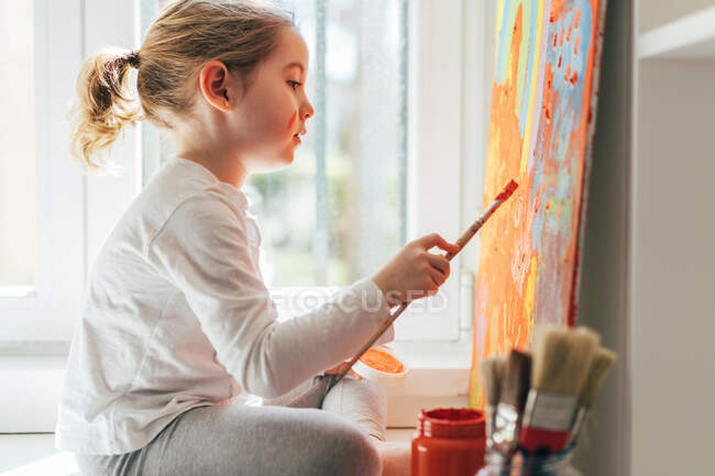Menina loira criativa em roupas casuais sentado no peitoril da janela contra janela e pintura com pincel grande arco-íris multi colorido na lona laranja — Fotografia de Stock