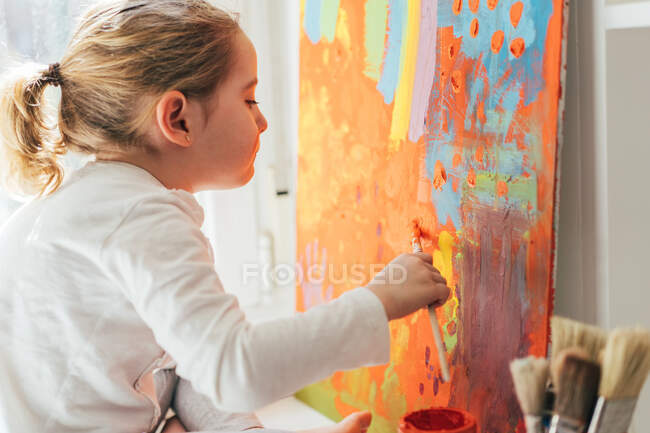 Fille blonde créative en vêtements décontractés assis sur le rebord de la fenêtre contre la fenêtre et la peinture avec pinceau grand arc-en-ciel multicolore sur toile orange — Photo de stock