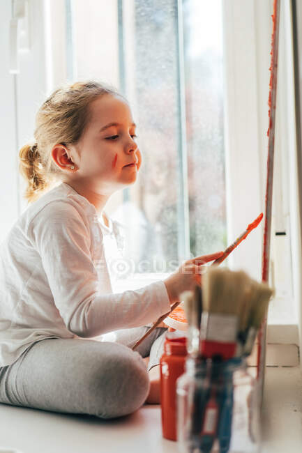 Menina loira criativa em roupas casuais sentado no peitoril da janela contra janela e pintura com pincel grande arco-íris multi colorido na lona laranja — Fotografia de Stock