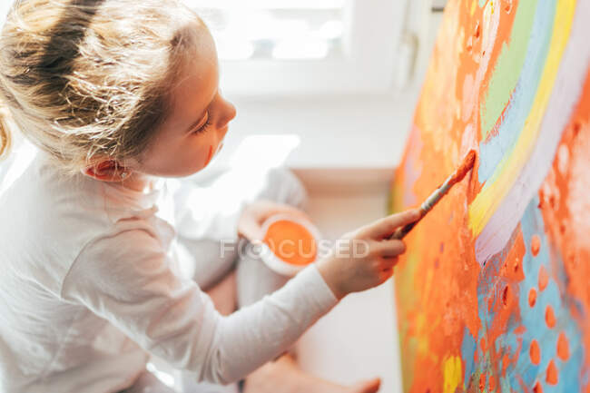 Chica rubia creativa en ropa casual sentado en el alféizar de la ventana contra la ventana y la pintura con pincel grande arco iris multicolor sobre lienzo naranja - foto de stock