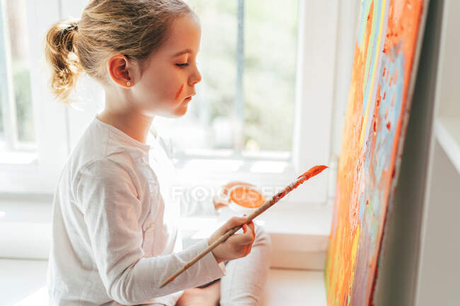 Creative blond girl in casual clothes sitting on window sill against window and painting with paintbrush large multi colored rainbow on orange canvas — Stock Photo
