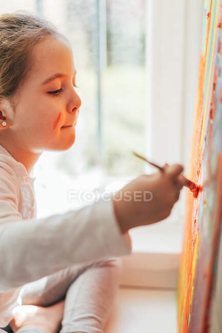 Творча блондинка у повсякденному одязі сидить на підвіконні біля вікна і малює з пензлем велику багатокольорову веселку на оранжевому полотні. — стокове фото