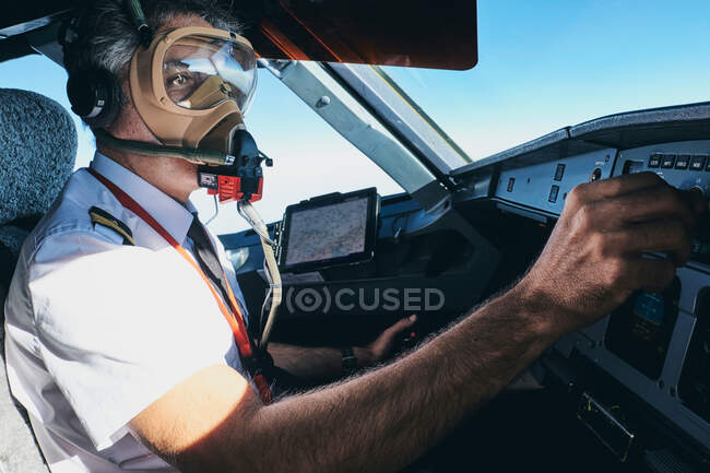 Side view of professional male aviator in oxygen mask turning switch on control console while operating  contemporary aircraft during flight — Stock Photo