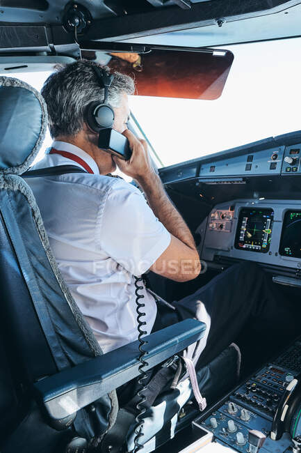 Seitenansicht des männlichen Kapitäns, der über UKW-Funk kommuniziert, während er im Cockpit moderner Flugzeuge sitzt und sich auf den Flug vorbereitet — Stockfoto