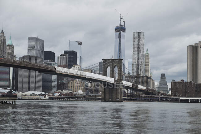 De dessous de Manhattan Bridge à partir de rive avec des bâtiments de la ville et un ciel gris nuageux en arrière-plan à New York — Photo de stock