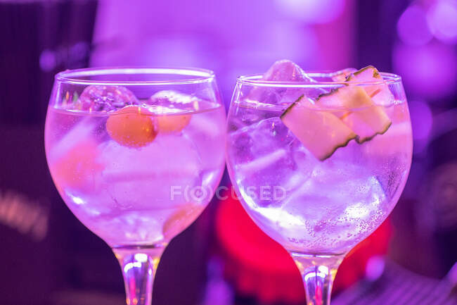 Glasses with fresh alcohol tonic cocktails with ice cubes and fruit served against blurred neon background — Stock Photo