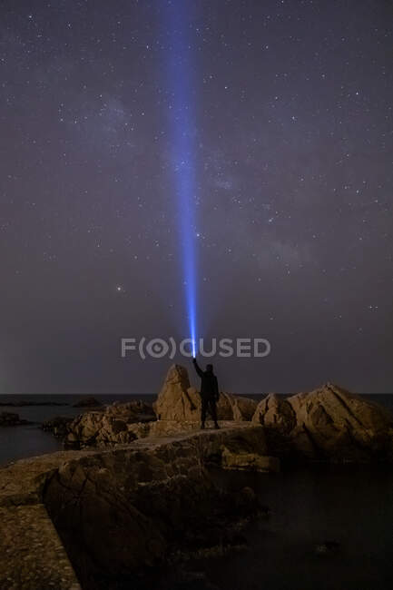 Silhouette of traveler with flashlight in raised hand standing on rocky coast with starry sky on background — Stock Photo