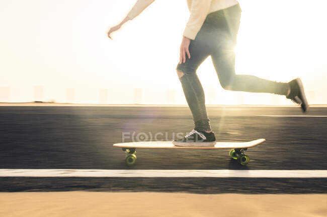 Unrecognizable guy in casual clothes riding skateboard on asphalt road in evening on Fuerteventura Island, Spain - foto de stock