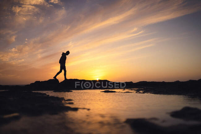 Silhueta de pessoa anônima andando na costa perto de água calma contra o céu do pôr-do-sol à noite na Ilha Fuerteventura, Espanha — Fotografia de Stock